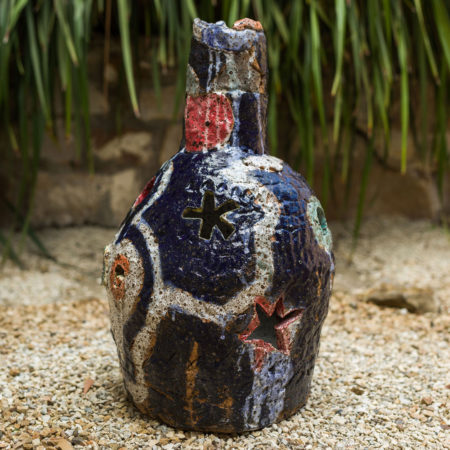 A large ceramic pot sits on a beige gravel surface with green foliage behind it. The bottle is mostly dark blue in colour with flashes of red, white and orange. There are star shapes cut out of the ceramic bottle shape.