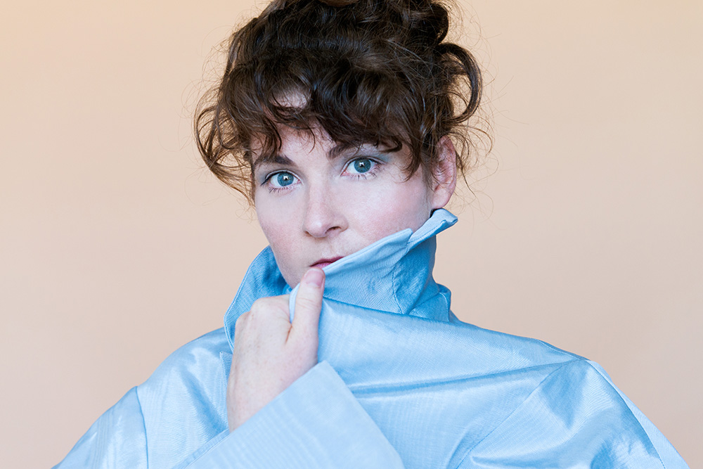 A white woman with brown hair and blue eyes looks directly to camera. She is wearing a blue silken top and has it covering part of her chin. We see her from the shoulders up. She stands in front of a peach background. 