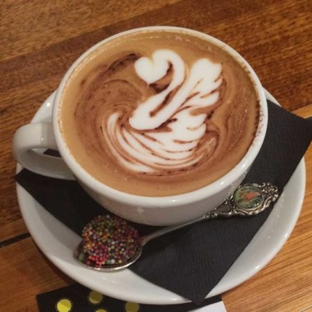 A photo of a frothy coffee on a saucer. A silver spoon lays across the saucer.