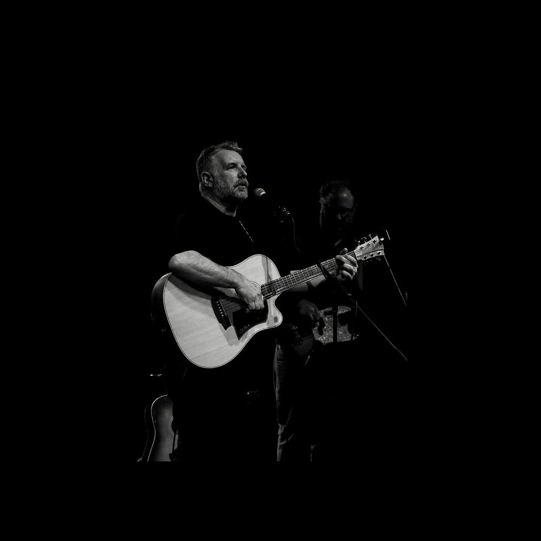 A man with greying hair plays a guitar on stage. The image is black and white.