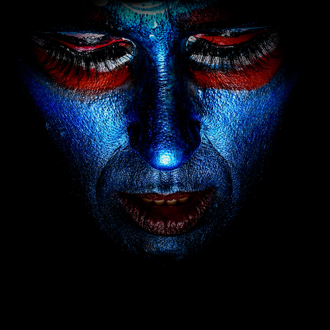 A woman's face is painted in iridescent blue and red to make her look like the Indian goddess, Kali. It is a close up of her face and she looks downward.