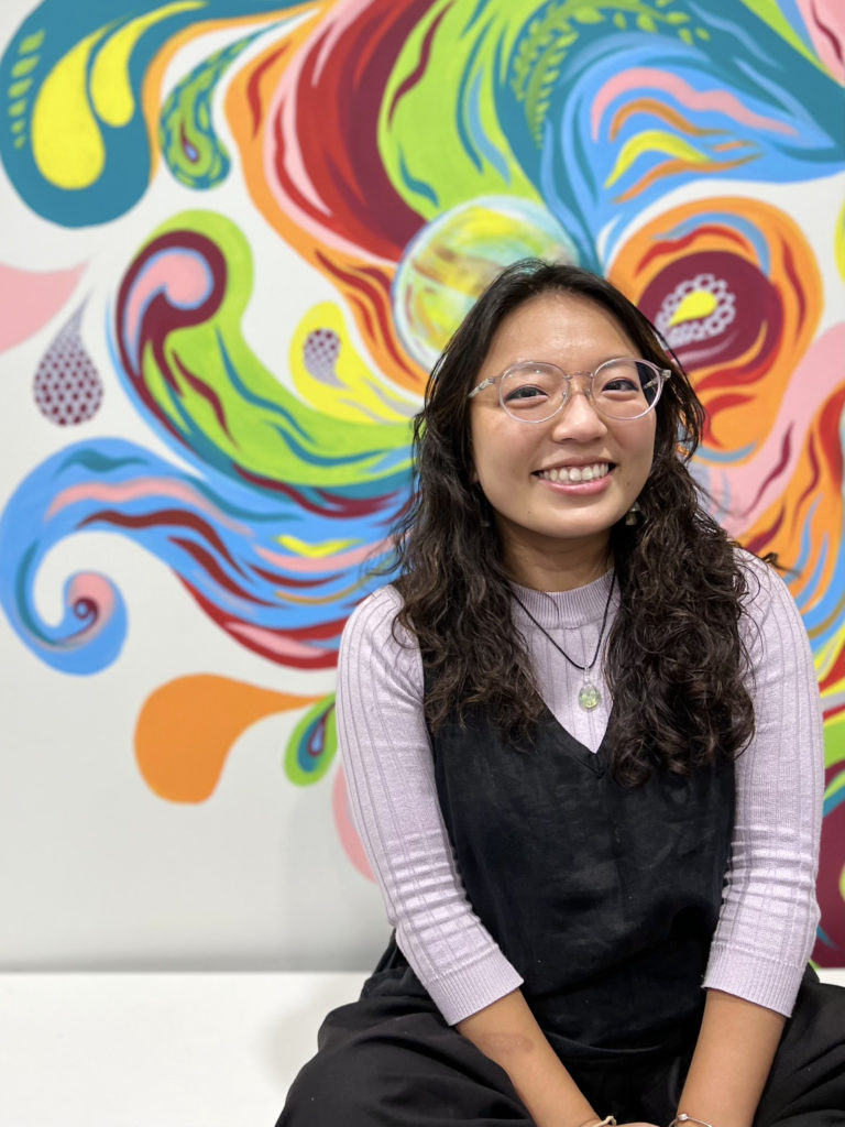 A japanese woman with long dark hair and glasses looks directly to camera, smiling. She sits in front of a wall with a coloured, swirly mural on it.