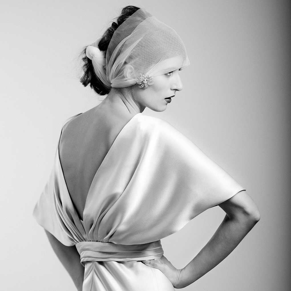 A black and white photograph of a woman wearing white flowing satin dress and a scarf wrapped around her hair.