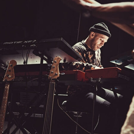 A man sits at the drums. He is white and wears a black beanie.