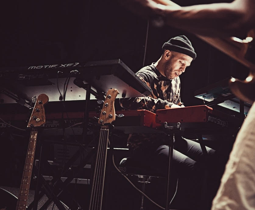 A man sits at the drums. He is white and wears a black beanie.