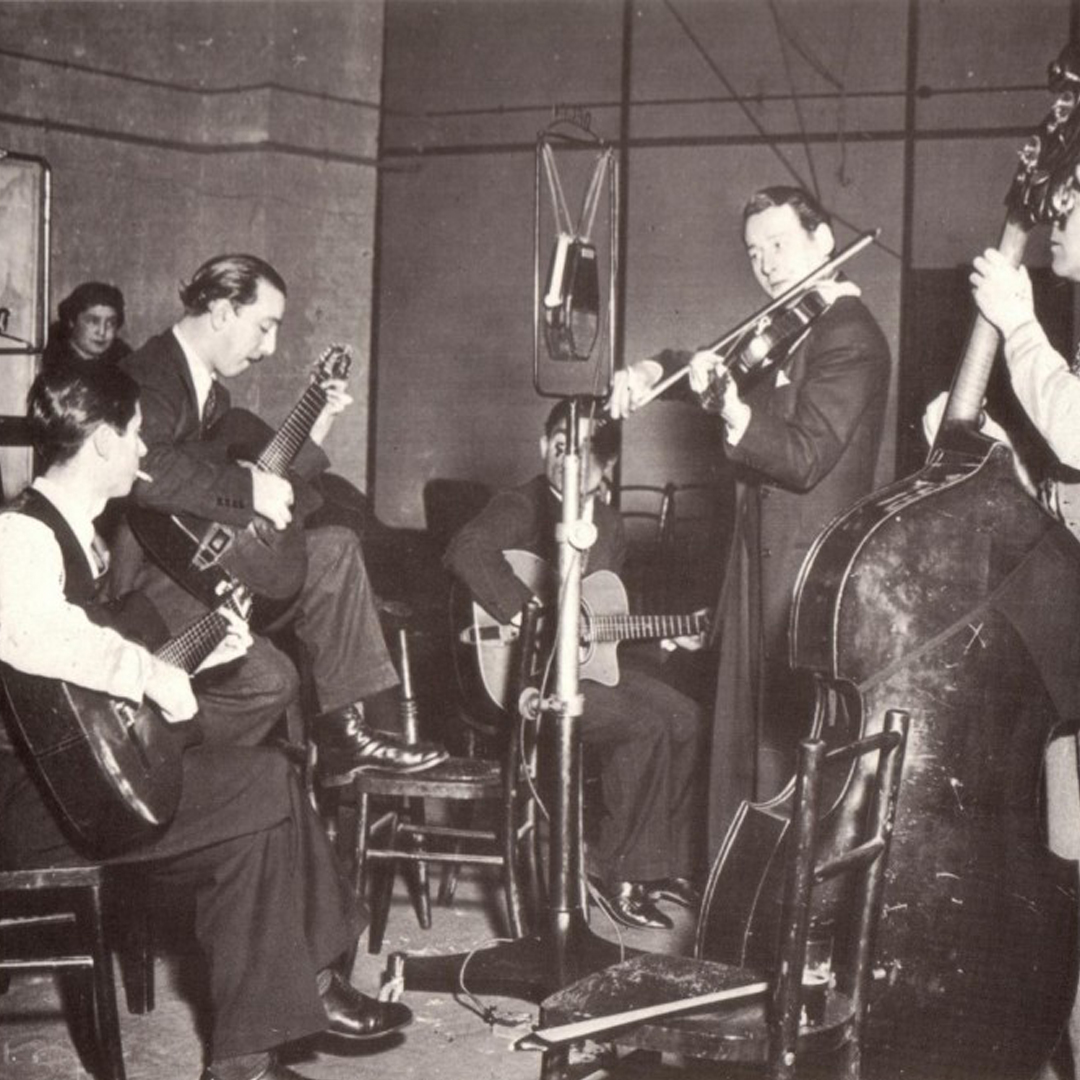 A black and white photo of a group of four, male musicians playing in the round. A guitarists is seated while the other three men stand.