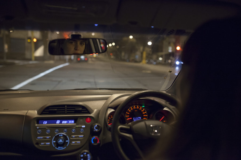 A woman is driving a car at night. We see the back of her head and her reflection in the rearview mirror