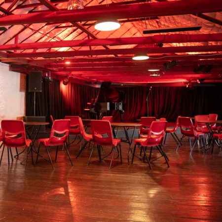 Photo pf the Founders Room : a room with dark wooden floors and exposed beams in the vaulted ceiling. In the foreground are red chairs and black tables set in cabaret configuraion. In the background is a stage with a baby grand piano aginst a black curtain backdrop.