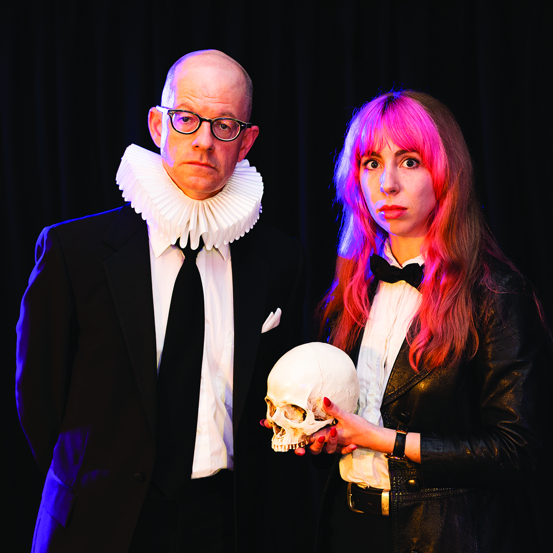 A bald, white man wearing dark rimmed glasses, a suit and a ruff around his neck. Standing next to him is a white woman with pink hair wearing a black suit and holding a human skull. The stand in front of a black background.