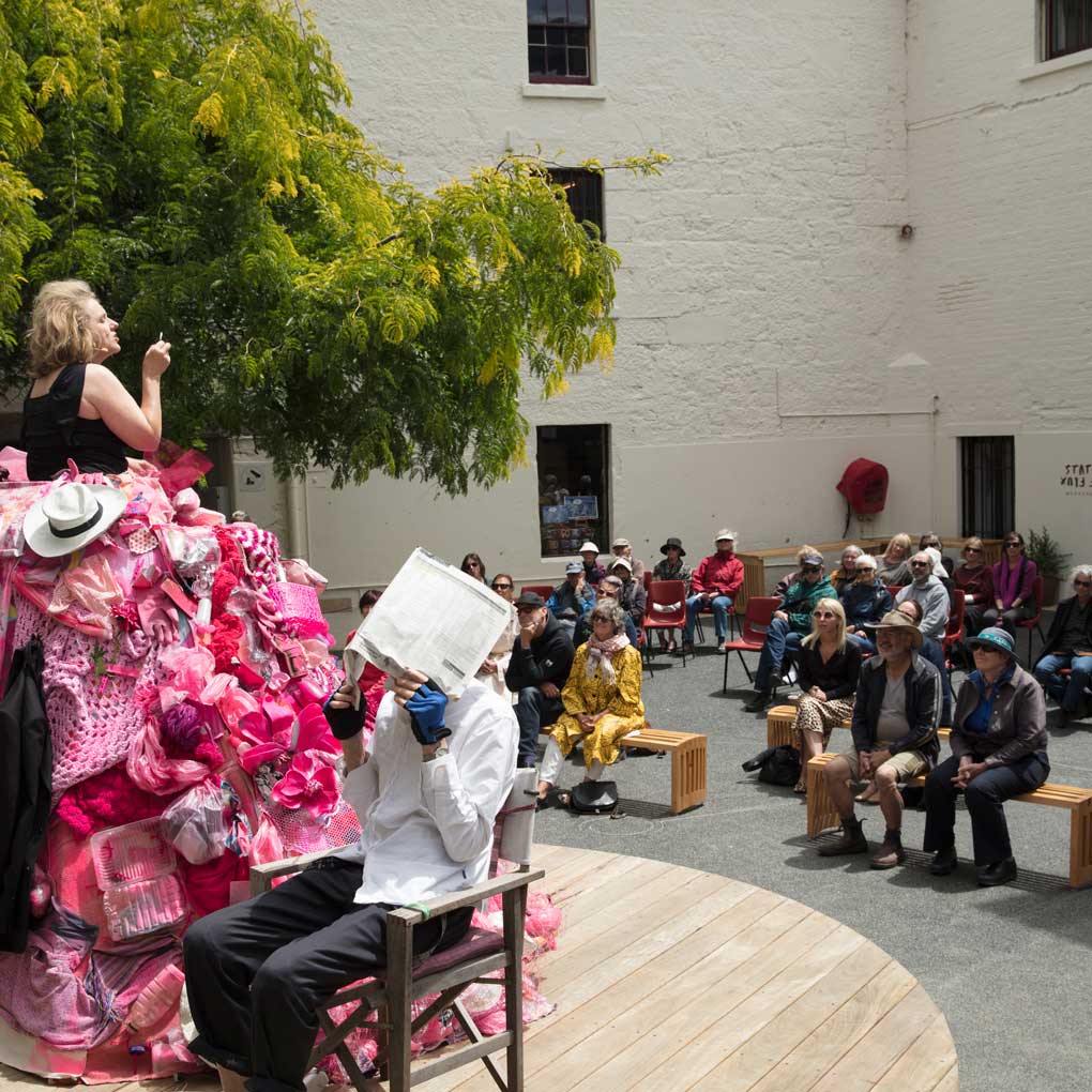 A performnce in the outdoor Courtyard : an actor is on stage wearing a large pink dress crraeted from various objects, whilst another actor on the stage sits in a chair facing away from the crowd. The crowd are seated, watching the performance.