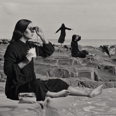 A woman sits in the foreground on a rock platform with binoculars. In the distance there are two other female figures, one seated looking out to see and one standing with arms outstretched looking towards the ocean. The image is black and white. The woman is Aboriginal and all three representations of her are wearing black clothing.