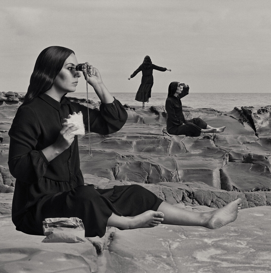 A woman sits in the foreground on a rock platform with binoculars. In the distance there are two other female figures, one seated looking out to see and one standing with arms outstretched looking towards the ocean. The image is black and white. The woman is Aboriginal and all three representations of her are wearing black clothing.