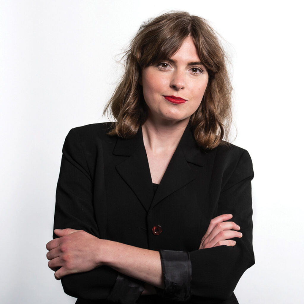 A white woman with shoulder length brown hair stands in front of a white background. She wears a black blazer and is wearing red lipstick.