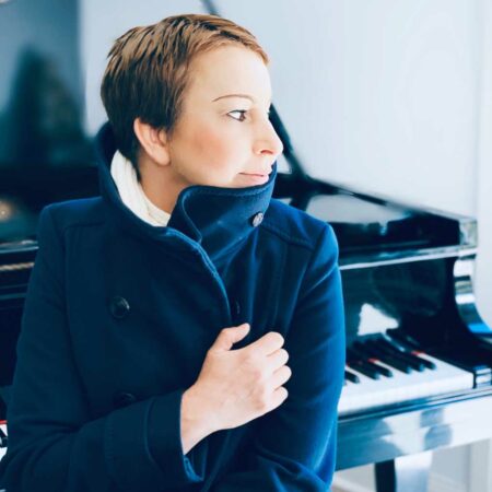 A woman with short hair with a dark coat wrapped tightly around her. In the background is a black piano.