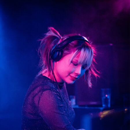 A DJ bathed in pink and purple light has headphones on and is looking downwards.