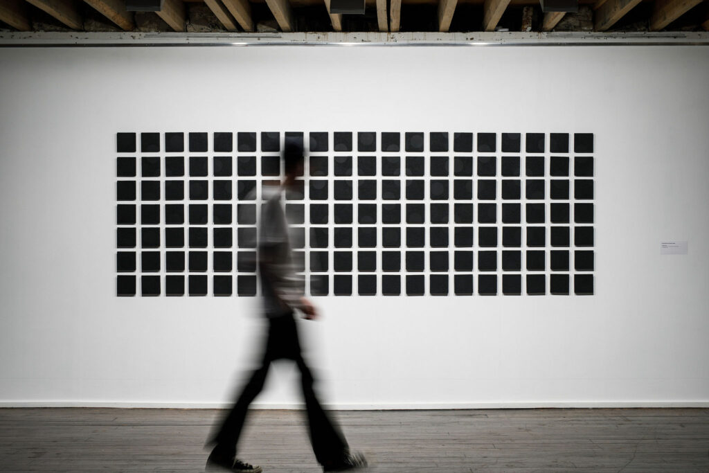 A grid of black square artworks sits on a white wall. A blurred figure passes in front of them. 