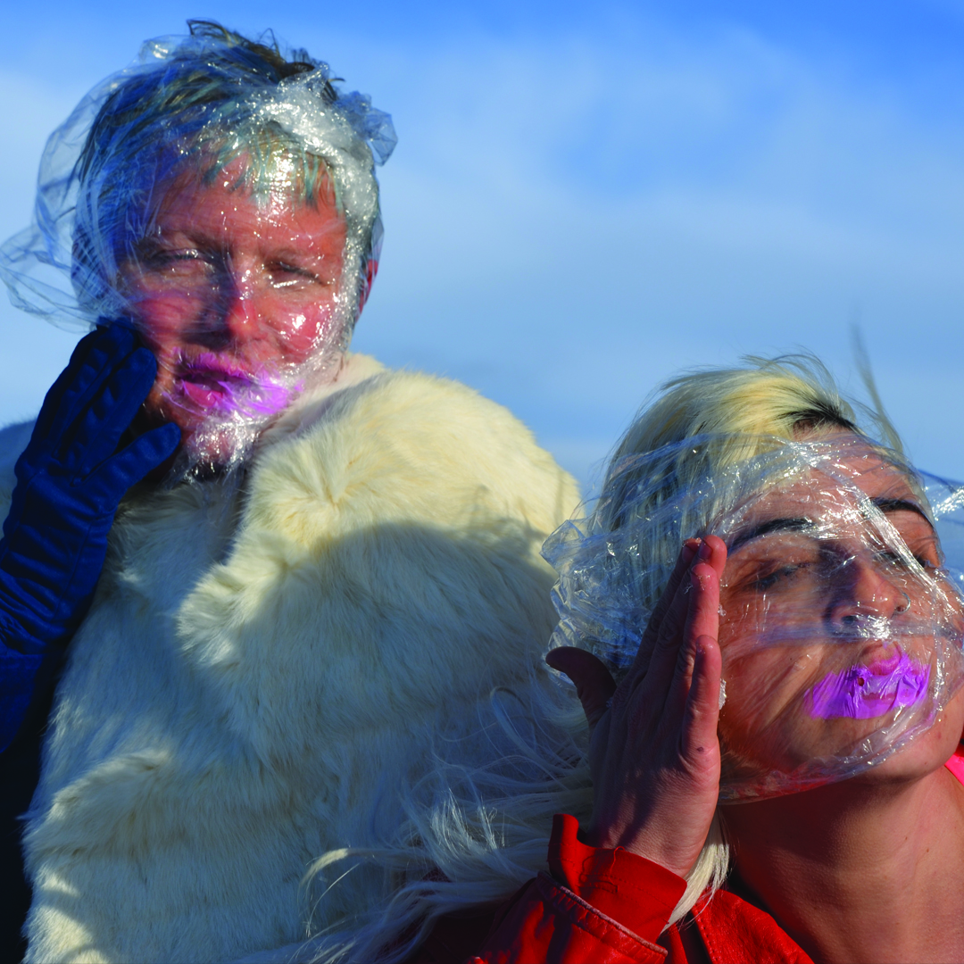 Two white women have their faces covered in cling wrap. They both wear pink lipstick which is smudged under the cling warp. Behind them is a blue sky. The woman on the left wears a white fur coat and the one on the right, has her hand to her face.