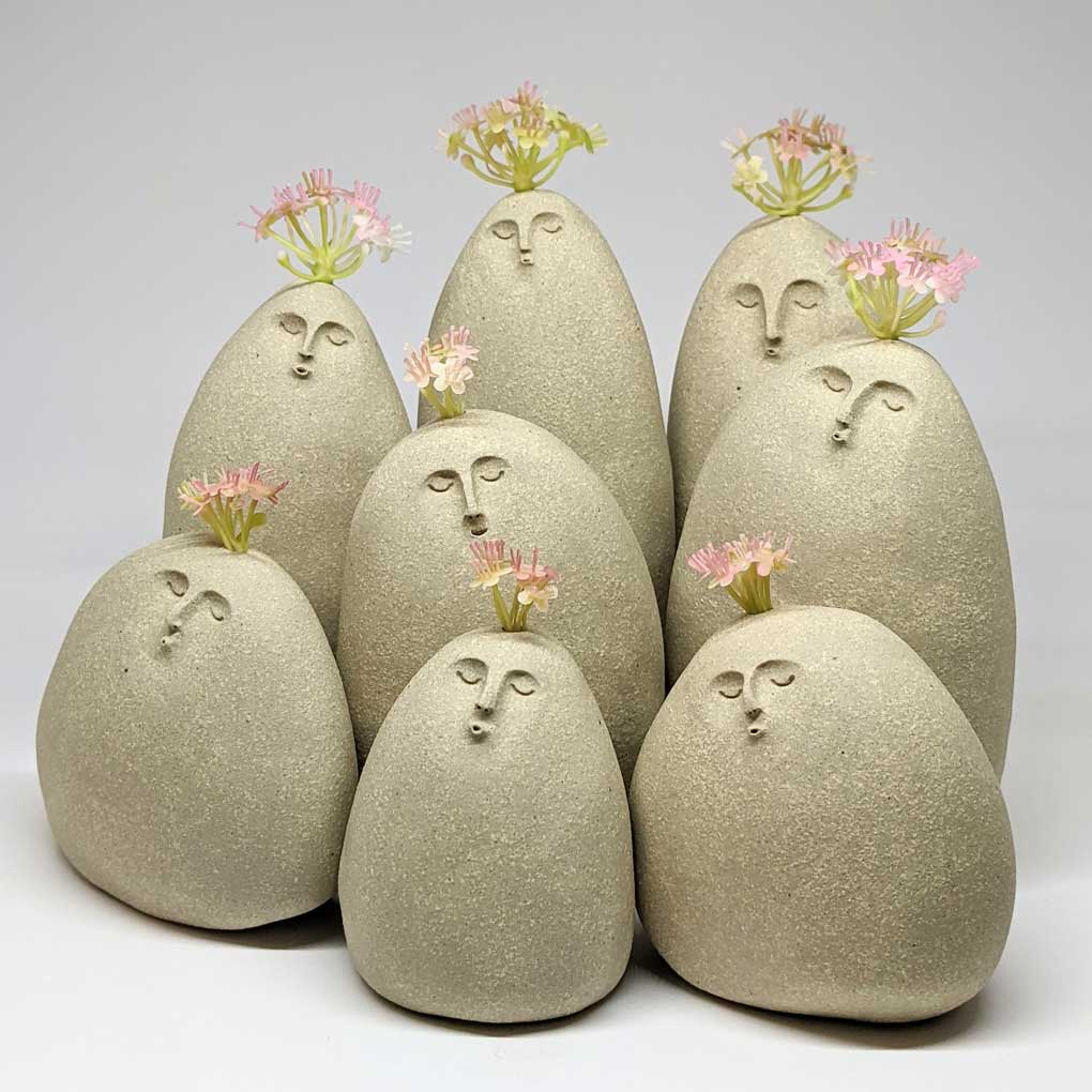 Eight, round, smooth, ceramic pebbles with faces. They have their eyes closed and their lips pursed as if whistling. Each has pink flowers sprouting from their head.
