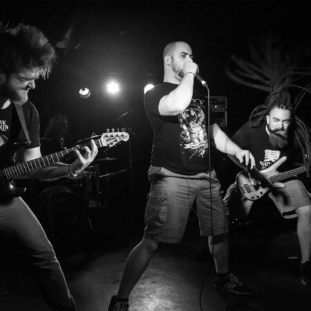 A black and white image of three men on stage. They are playing metal music and are really getting into it.