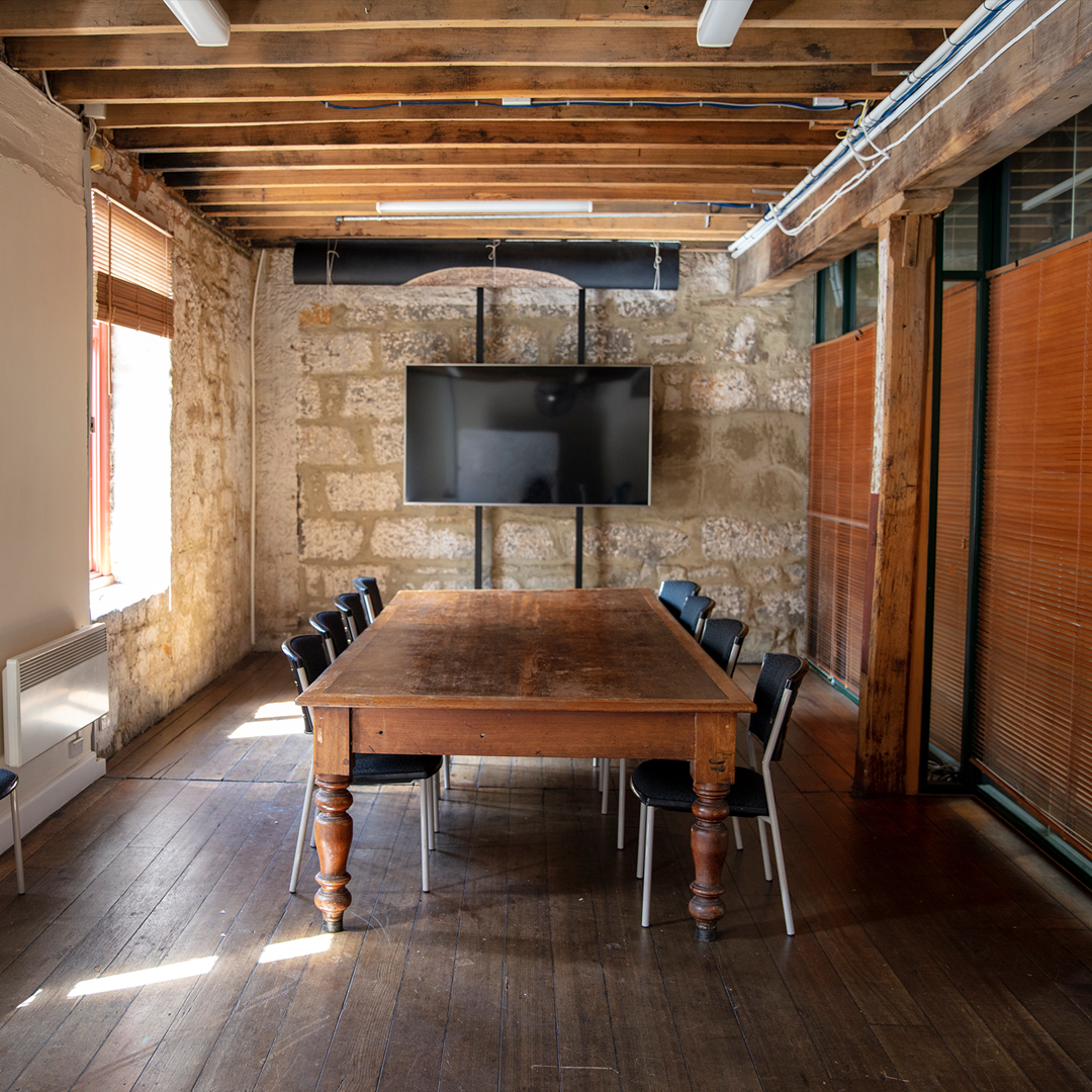 Photo of the Meeting Room : A large flat screen tlevision is mounted on the sandstone wall, whilst on the other wall there are wooden blids. The corner of an antiqu wooden tablr is visible, with blue chairs at the table.