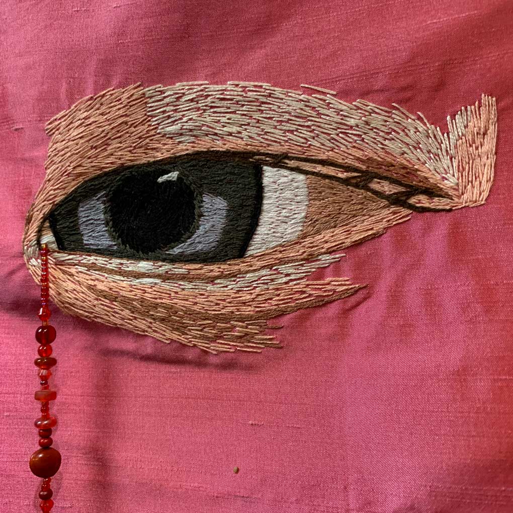 A grey eye embroidered on a pink cloth, with a string of red beads suspended from the tear duct.