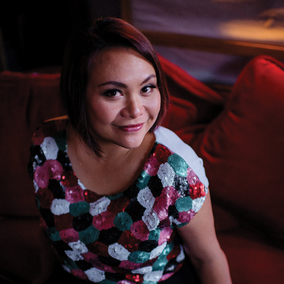 A woman looks directly to camera. She wears a multicoloured top and has a short, red bobbed haircut.