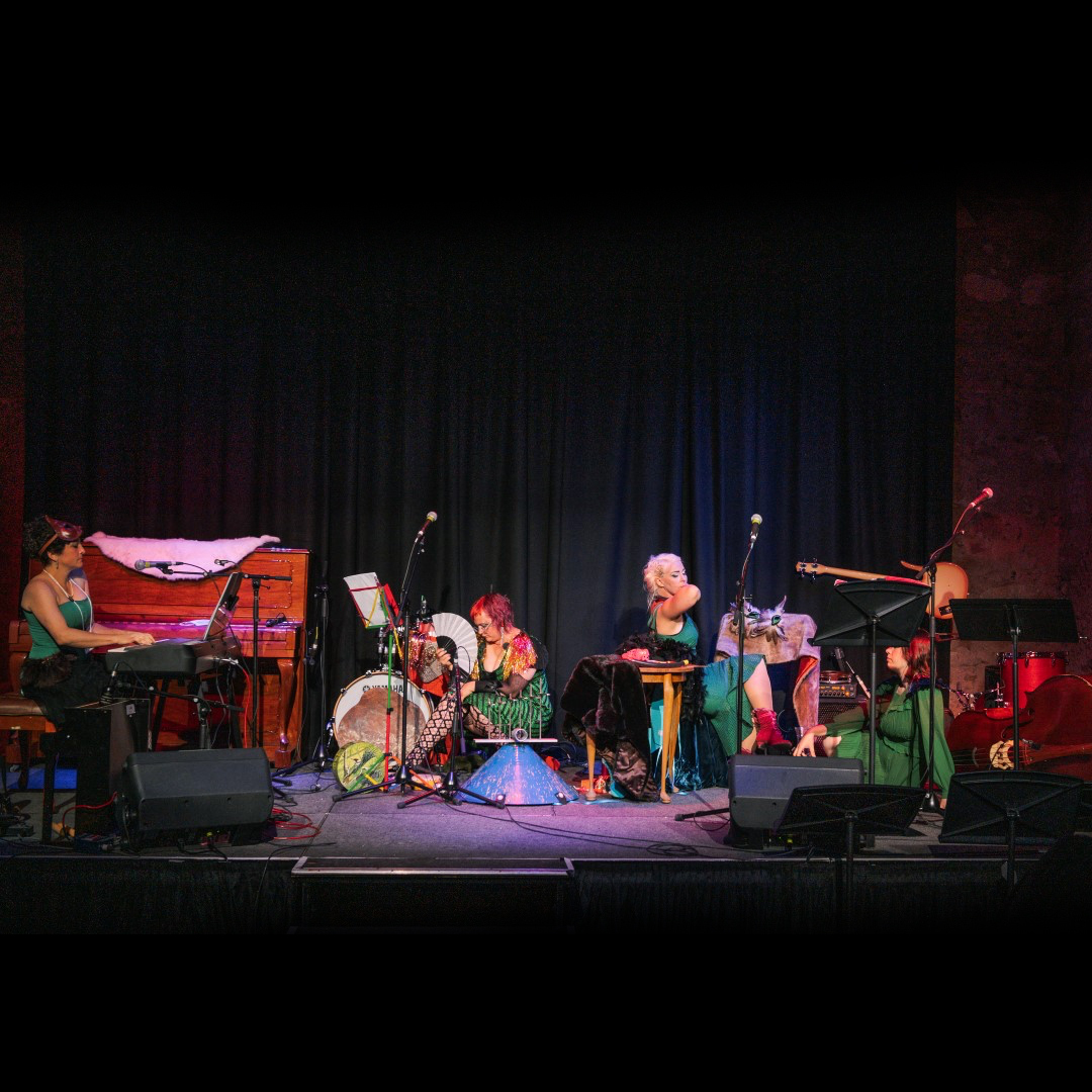 A group of all female performers sit on a darkened stage. We see them from a distance.