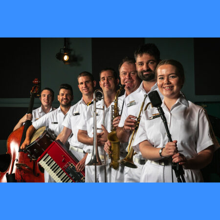 The Royal Australian Navy Band Melbourne Jazz Ensemble pose for a 2022 promotional photo at HMAS Cerberus, Victoria. A group of musician wearing the white navy uniform and holding their instruments stand in a line facing the camera. They are all smiling.