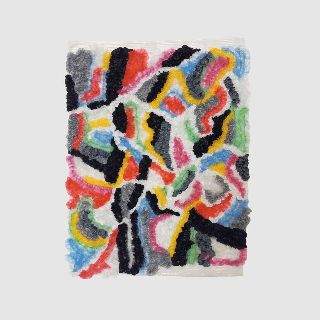 A swirling, colourful abstract pattern created from the weaving and knotting of found, soft plastics. It sits on a blank, white wall.