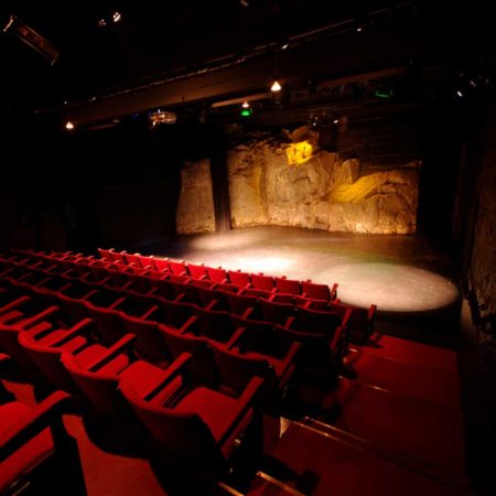 Photo of the Peacock Theatre. Red rows of theatre seats in the foreground, looking towards the stage. The stone wall of the back of the stage is lit with warm coloured theatre lights.
