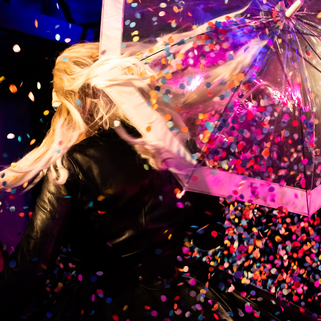 An image of someone with long blond hair. They are turned away from the viewer. They hold a clear umbrella over their head. Confetti is being thrown.