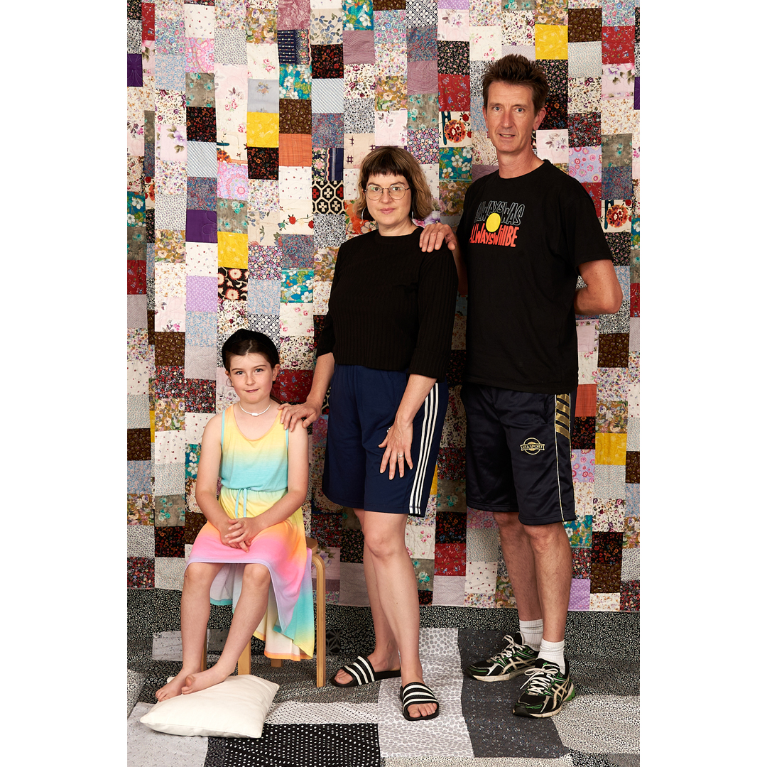 A family of three, stands in front of a patchwork quilt of may colours. The family is made up of a woman, a man and a girl. The girl is seated and approximately 8 years old. She wears a gradient coloured dress in tropical colours. The mother stands next to her with her left hand on her shoulder. The mother has shoulder length bob and wears glasses. The man at the right of the image has his left hand on the mother's right shoulder. They all look a bit awkward.