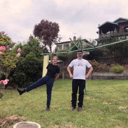 A man and woman stand in a grassy backyard. The woman has one of her legs off the ground. The main has his hands on his hips. They are looking towards the camera.