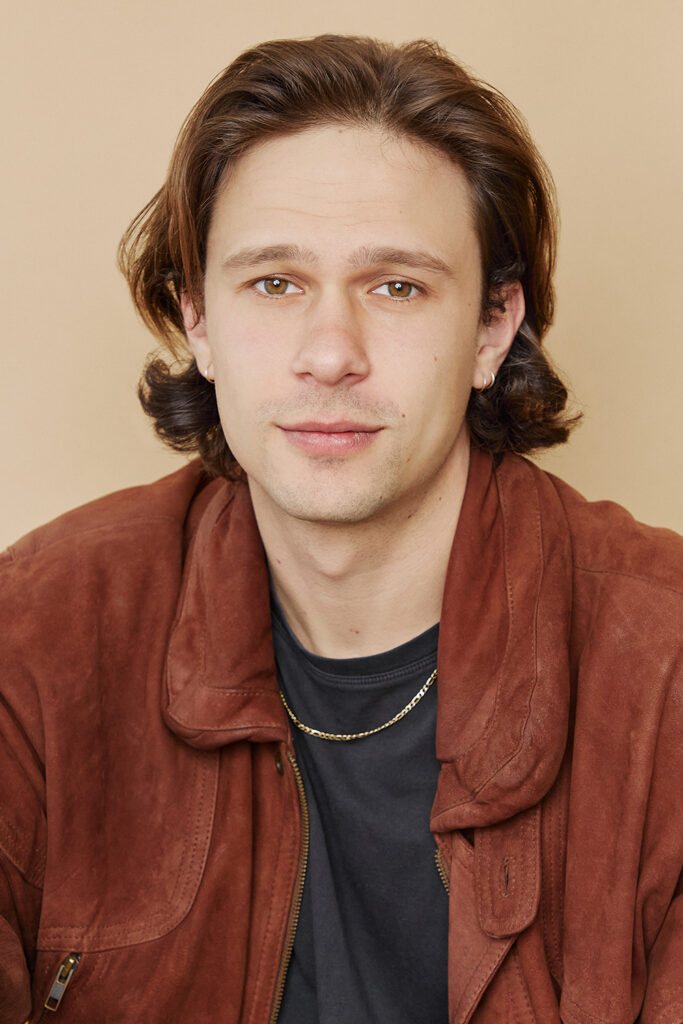A white male looks towards the camera. He wears a brown jacket, a black teeshirt and a gold chain. He has long-ish brown hair to his shoulders and light green eyes. 
