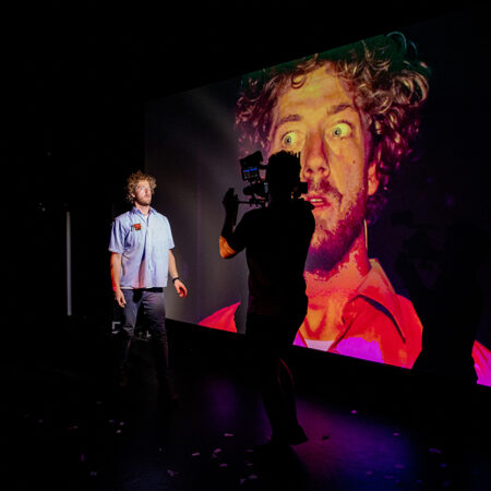 A young, white male stands in front of a person with a video camera. The video camera image is projected in an image behind the man and is large and filtered red. The man's eyes are wide and he has blond curly hair.