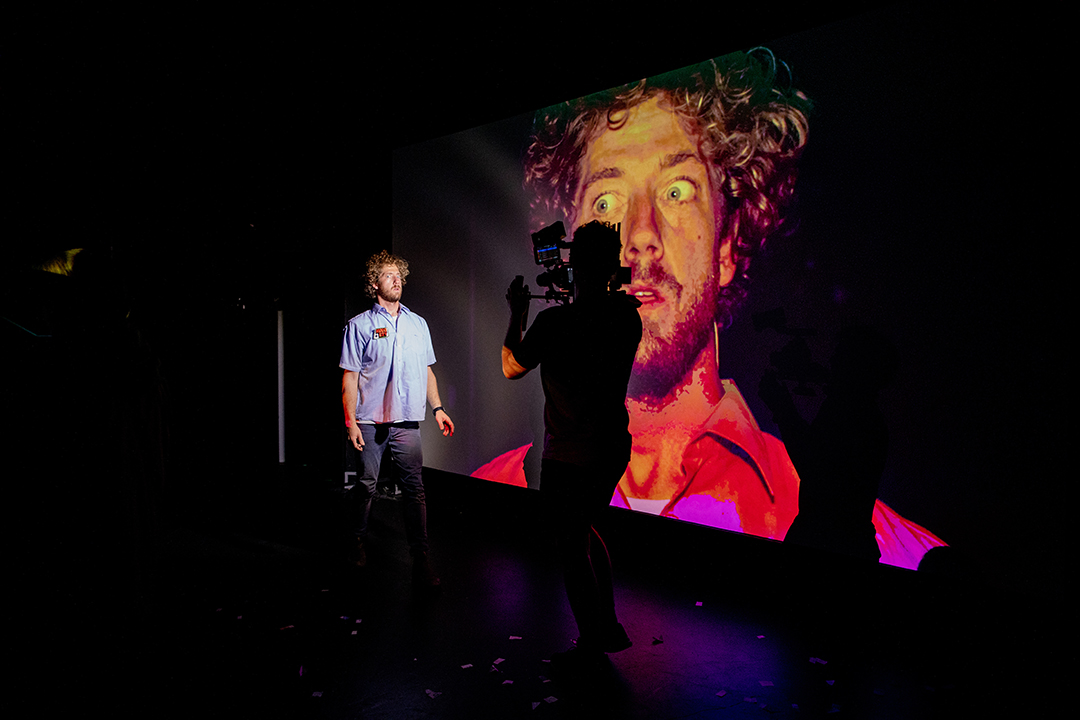 A young, white male stands in front of a person with a video camera. The video camera image is projected in an image behind the man and is large and filtered red. The man's eyes are wide and he has blond curly hair.