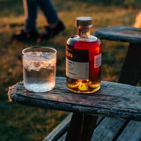A photograph of a bottle of whisky and glass of whisky balanced on the arm of a wooden outdoor chair. In the background a persons feet and visible as they walk past.