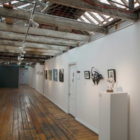 Photograph of the Studio Gallery : A long room / hallway with many doors leading off it and small framed artwork hung on the wall.The wall on the right is white and the far wall is grey. Low beams span the gallery walls and dark wooden floor.