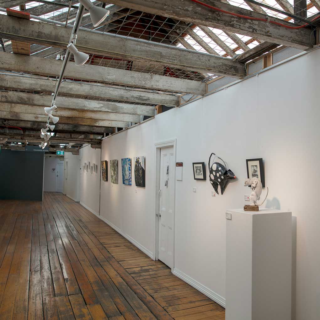 Photograph of the Studio Gallery : A long room / hallway with many doors leading off it and small framed artwork hung on the wall.The wall on the right is white and the far wall is grey. Low beams span the gallery walls and dark wooden floor.