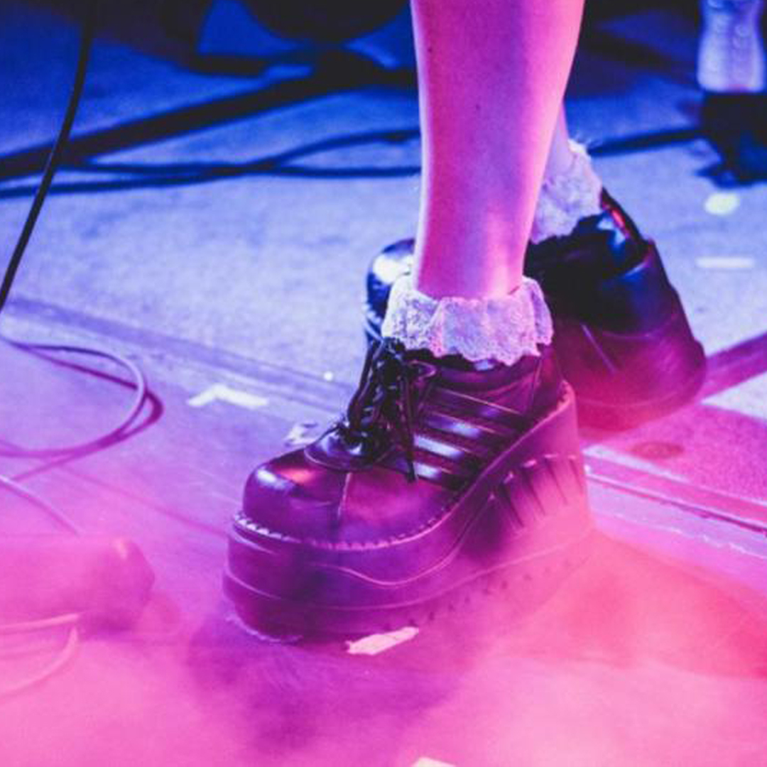 A pink, purple and blue gradient image of a pair of feet in chunky black shoes. They are wearing white, frilly socks. There looks to be microphone leads in the background of the image on the floor.