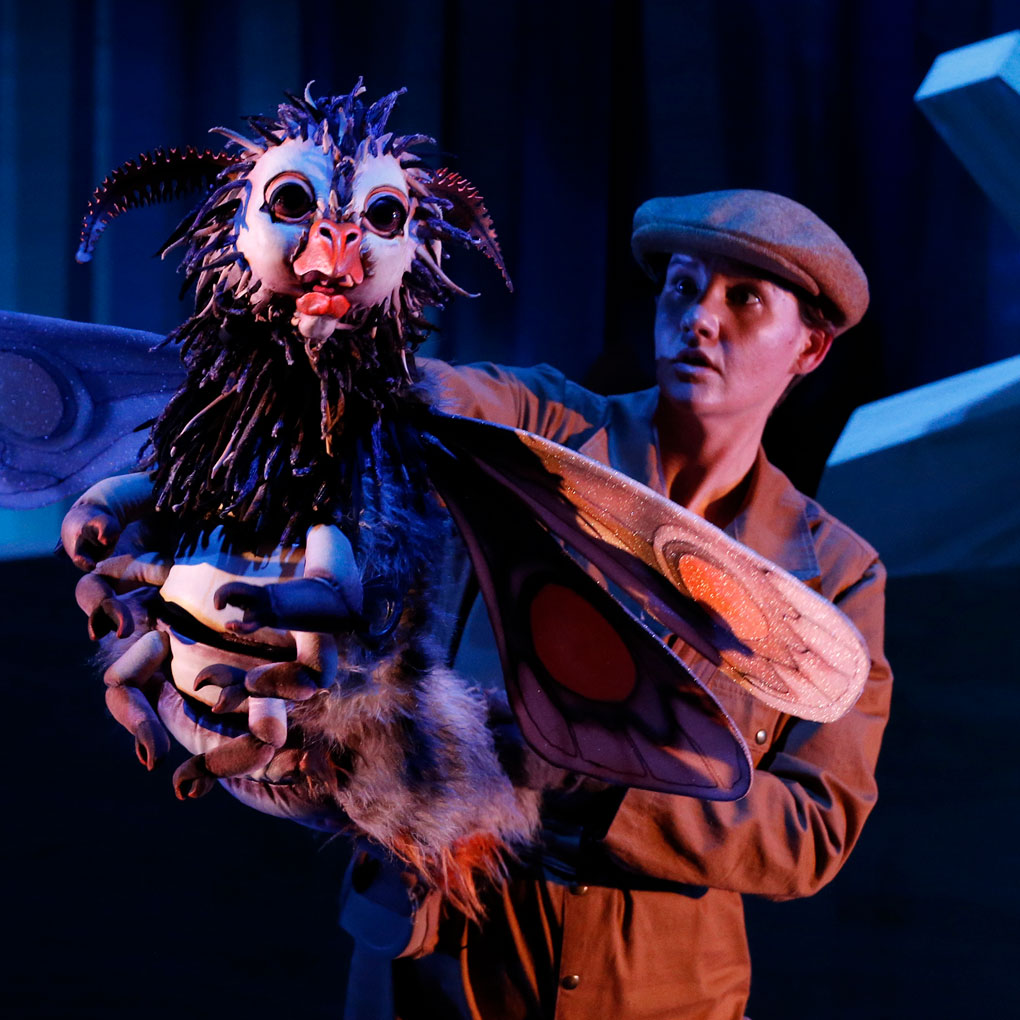 A puppeteer wearing a newsboy cap holds an oversized creature puppet