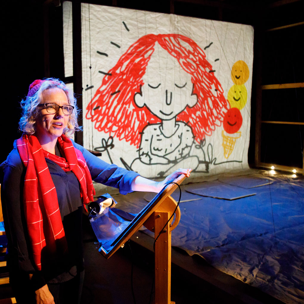 A woman in a red scarf and beanie stands at a podium. Behind her is a large cartoon drawing of a girl with bushy red hair and fuzzy jumper.