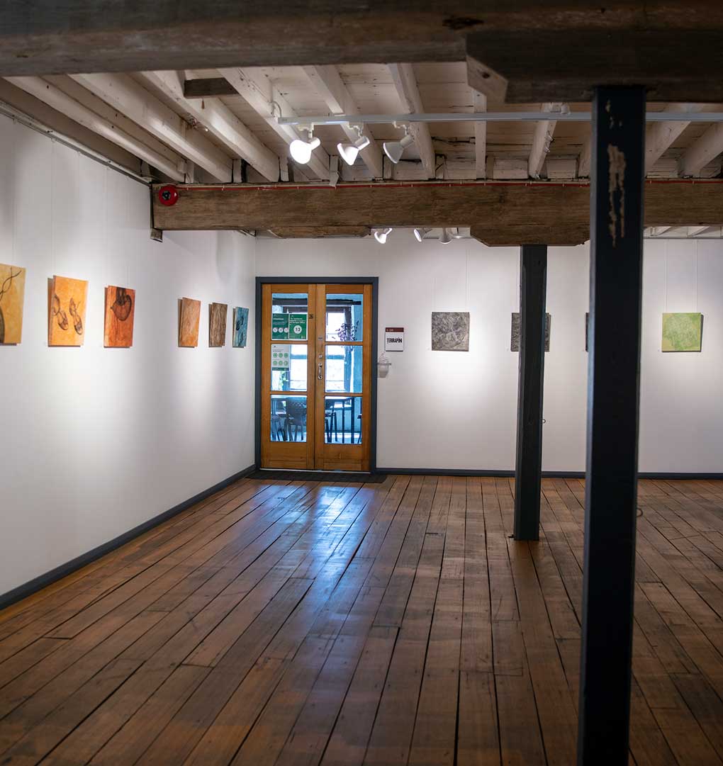 Photograph of the Top Gallery : A room with white walls and dark wooden floor. Two support beams are in the middle of the room. To the far end of the room is a wooden, french multi-paned door.