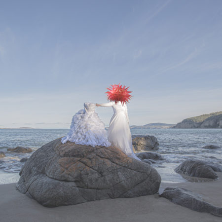 A woman with a giant sea anenemy stands on a large rock by the sea. She figure wears all white and holds up a white piece of fabric to her left hand side.