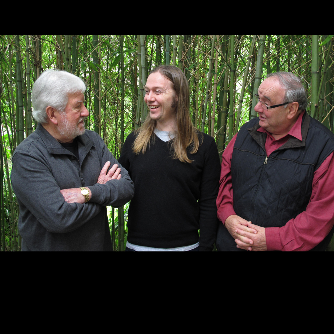 three men stand in front of a bamboo forest. The man on the left has grey hair and looks to the long haired man in the middle. The man on the right wears glasses and looks to the man in the middle also.