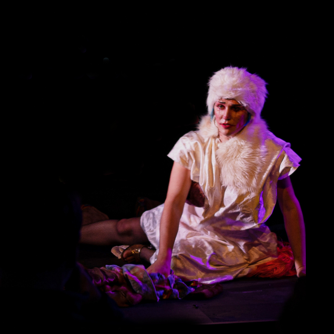 A performer on stage wearing white clothing and a white fluffy hat looks out to the audience with a forlorn look. The lighting on them is red and purple.