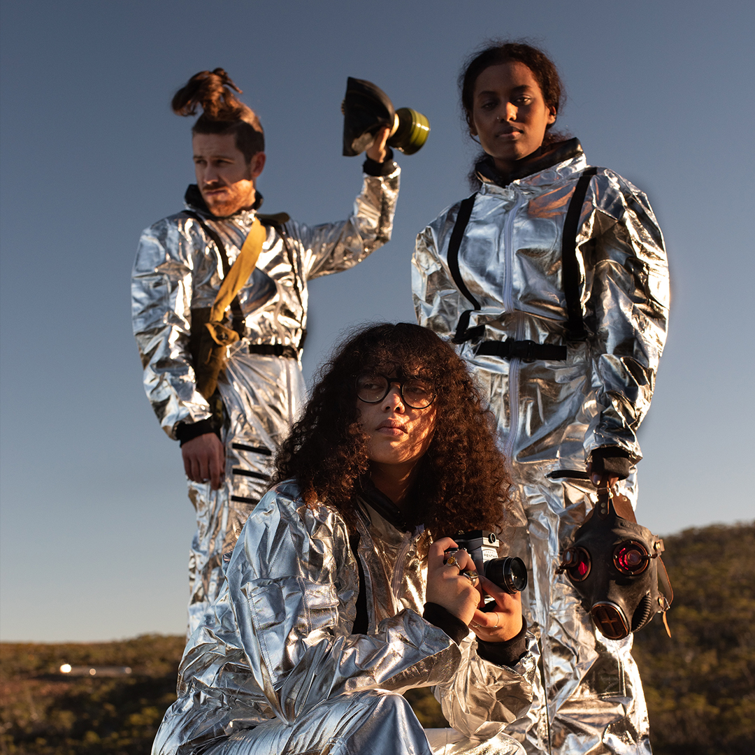 A group of three people wearing silver suits are on top of a mountain. The figure in the front is crouched down and has shoulder length curly hair and wears glasses. The firgure standing on the left holds a gasmask in their hand. The figure on the left has their hair in a top knot poinytail and holds something up in their left hand.