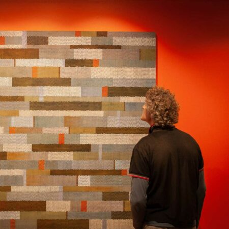 A person looking at an artwork hanging on the wall. The artwork is made of squares of natural fibre against an orange wall.