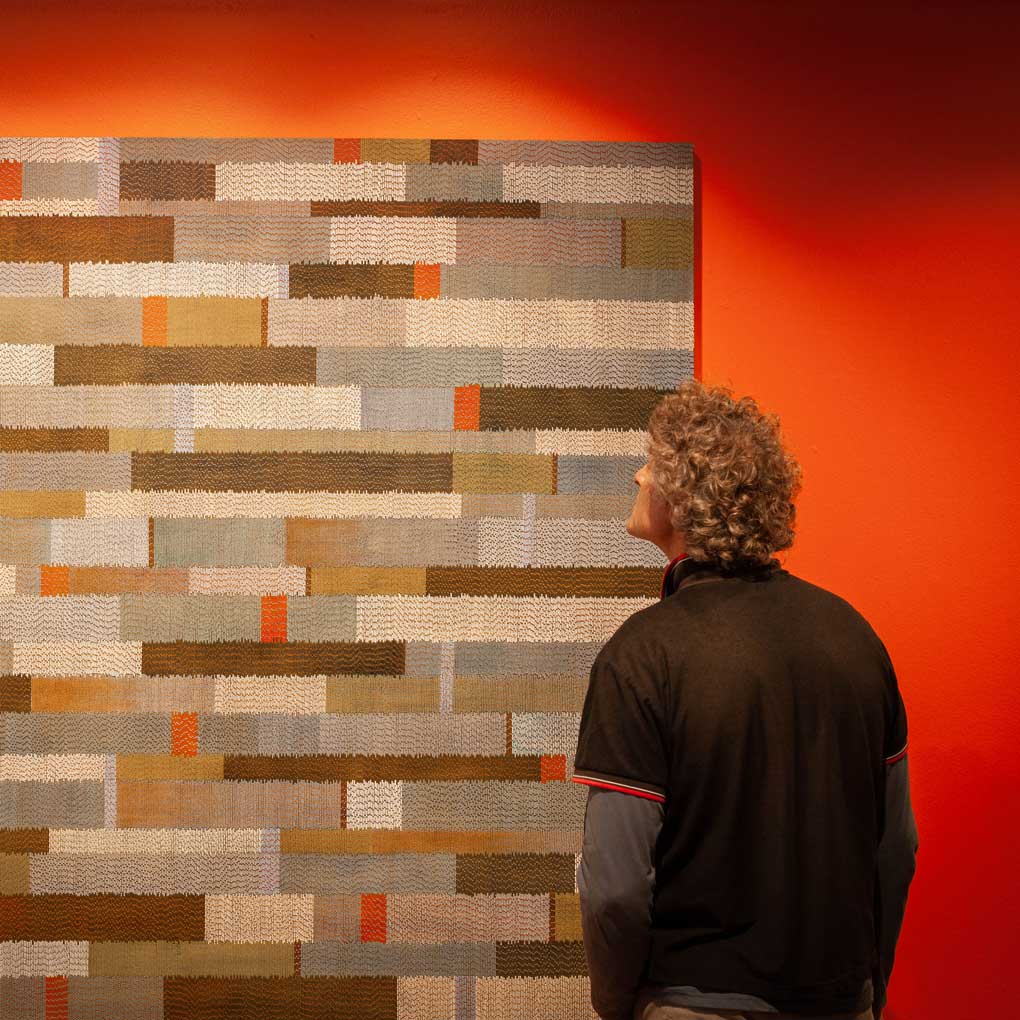 A person looking at an artwork hanging on the wall. The artwork is made of squares of natural fibre against an orange wall.