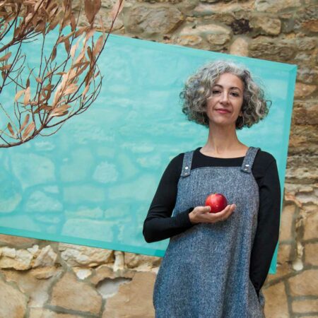 A woman in a black long sleeve top and blue apron dress, holding a red apple. In the background is a textures sandstone wall, which is partially covered by a square of semi-transparent light blue fabric. There is a dry tree branch in the corner.